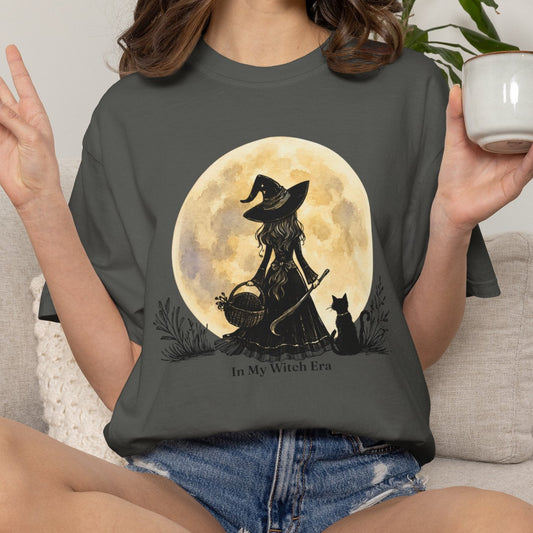 A woman sitting on a light beige couch wearing a dark gray graphic t-shirt with a witch silhouette design in front of a large moon, holding a white mug.