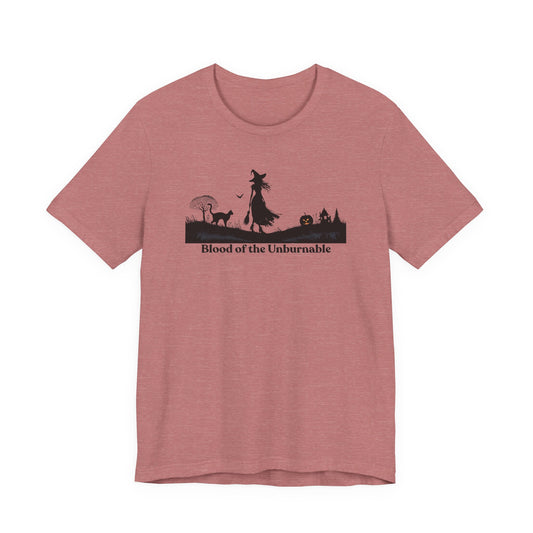 Pink t-shirt featuring a black silhouette design of a witch with flowing hair and broom, a black cat, a pumpkin with a glowing face, and a spooky house in the background, with the phrase “Blood of the Unburnable” printed below.