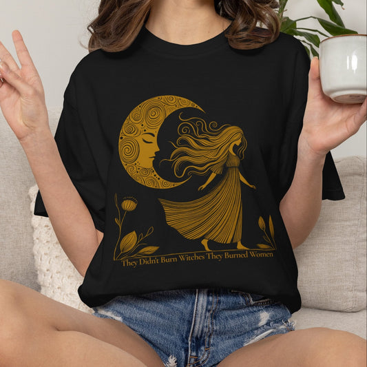 A woman sitting on a light beige couch wearing a black graphic t-shirt with a gold crescent moon and woman design, holding a white mug.