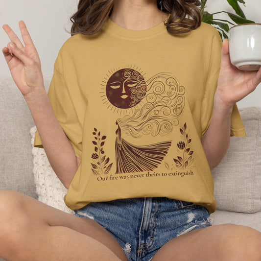 A woman sitting on a light gray couch wearing a mustard yellow graphic t-shirt with a brown sun and woman design, holding a white mug.