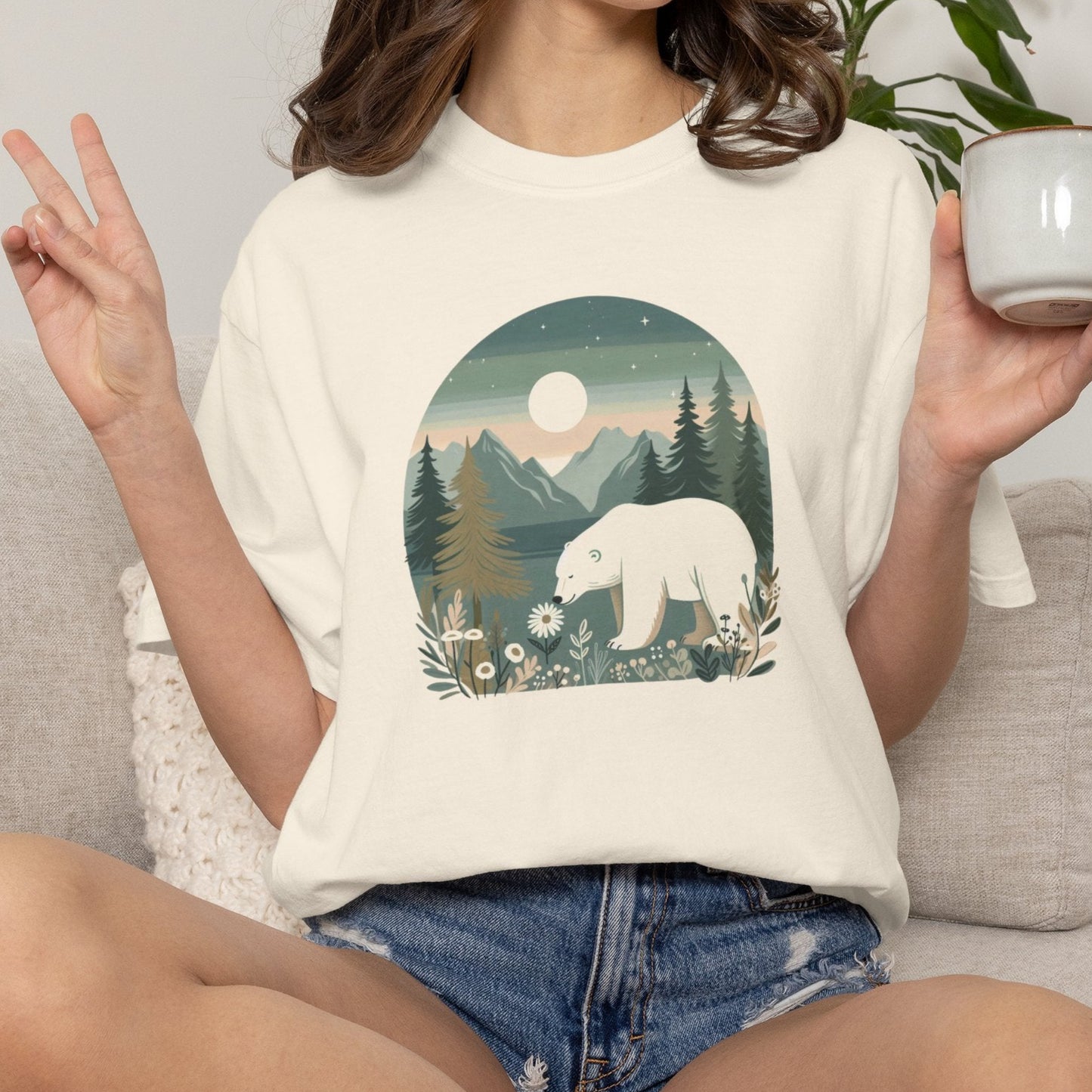 A woman sitting on a light gray couch wearing a cream-colored t-shirt with a graphic print of a polar bear in a forest at night. She is holding a white ceramic mug in her hand, with a potted plant in the background.