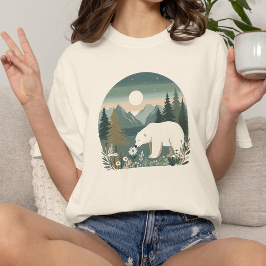 A woman sitting on a light gray couch wearing a cream-colored t-shirt with a graphic print of a polar bear in a forest at night. She is holding a white ceramic mug in her hand, with a potted plant in the background.