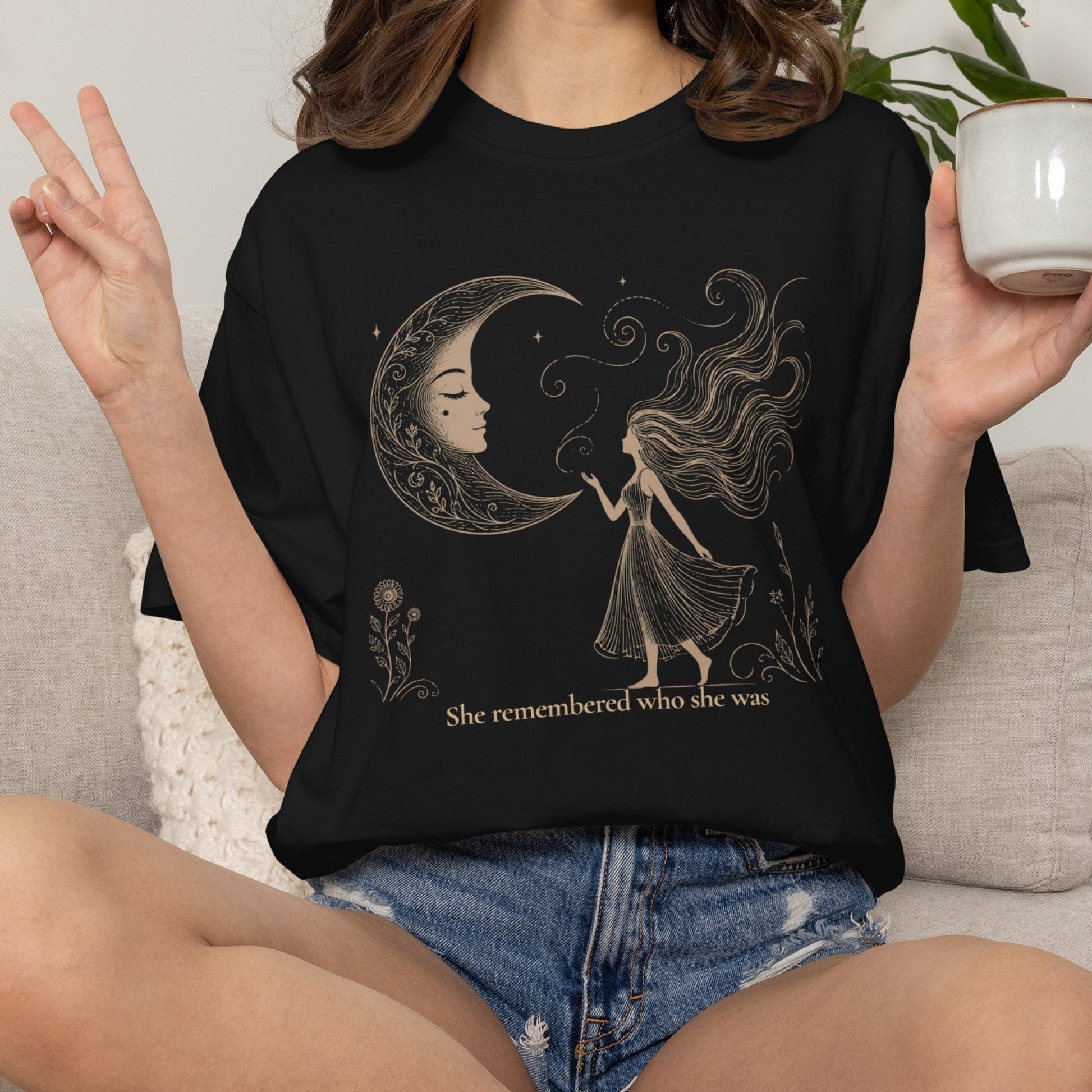A woman sitting on a light beige couch wearing a black graphic t-shirt with a gold-toned celestial design of a crescent moon and a girl, with the text 'She remembered who she was'. She is holding a white mug.